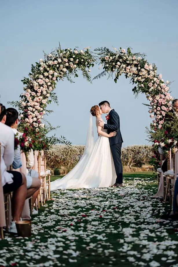 Bride and groom fist kiss under a floral wedding arbor - Madiow Photography
