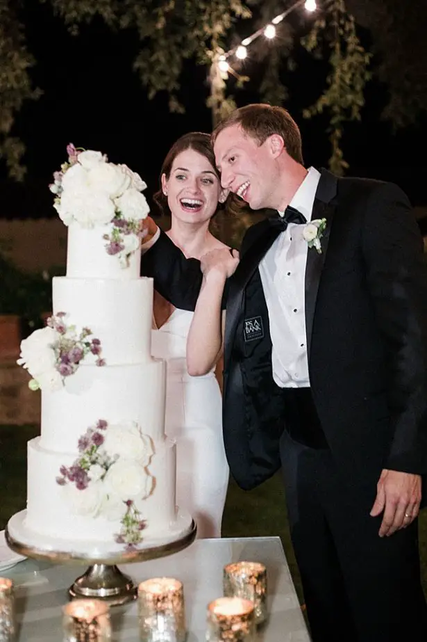 Bride and groom cutting four tiered all white wedding cake with white and mauve flowers - Purewhite Photography