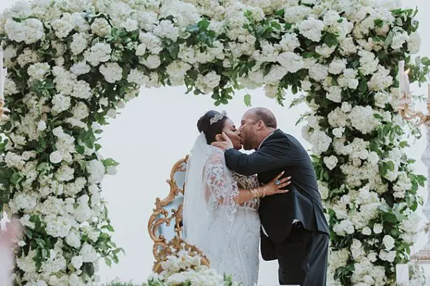 Epic photo of bride and groom first kiss with floral arch behind them - Photo: Dmitry Shumanev Production